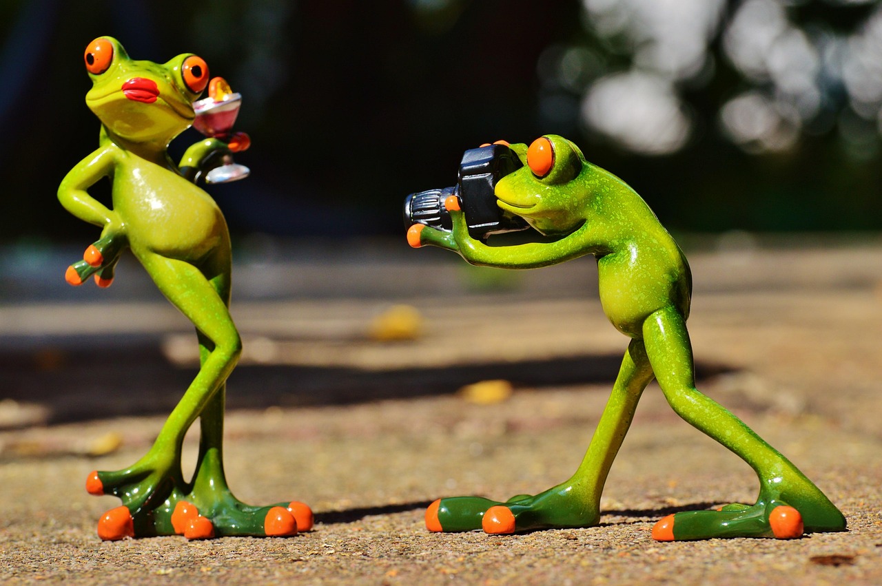 Two green frog figurines staging a playful photoshoot—one posing with a cocktail, the other holding a camera.